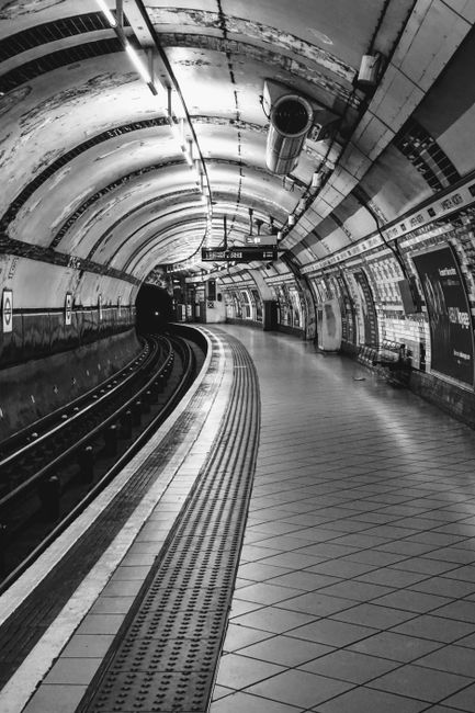 A black and white photograph of an underground Tube station tunnel with curved walls and ceiling, featuring tiled flooring and tactile paving along the platform edge. The station walls are lined with 